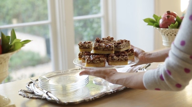 a person serving pecan bars on a cake stand and placing them on a silver tray set with glass plates
