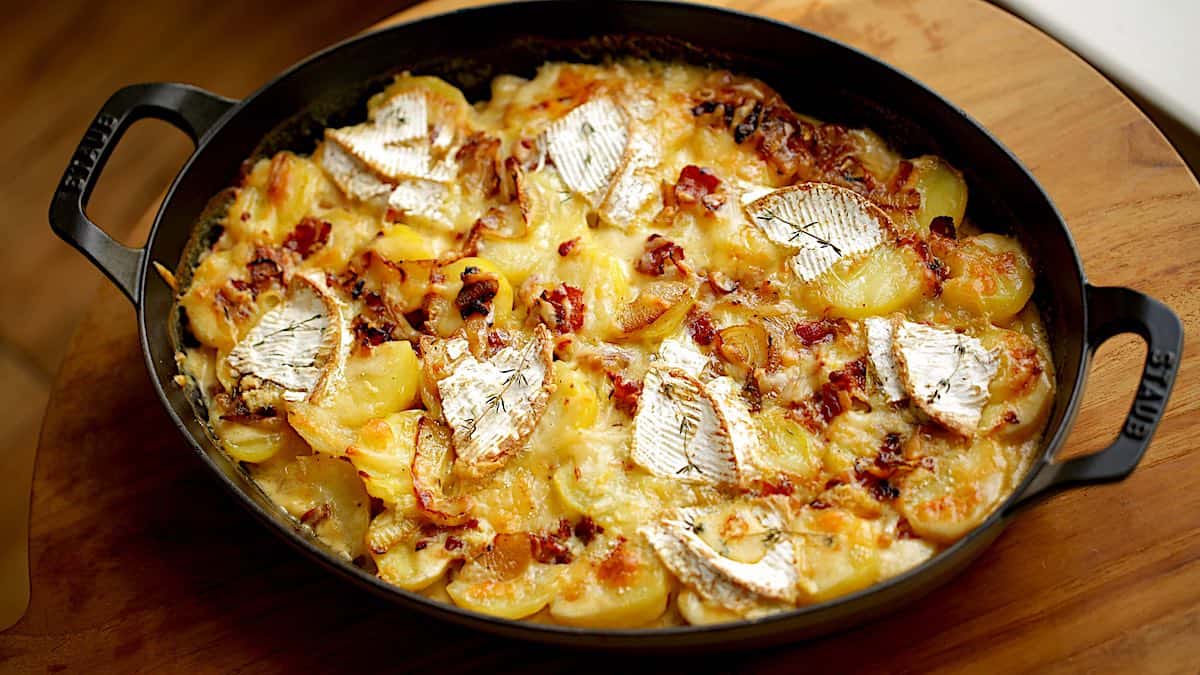a gratin dish filled with sliced potatoes, bacon, and brie cheese