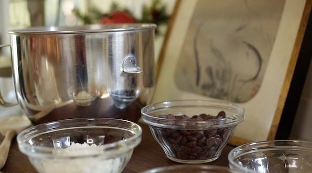 Bowls of chocolate chips, sweetened coconut, nuts and cornflakes going into a bowl of an electric mixer