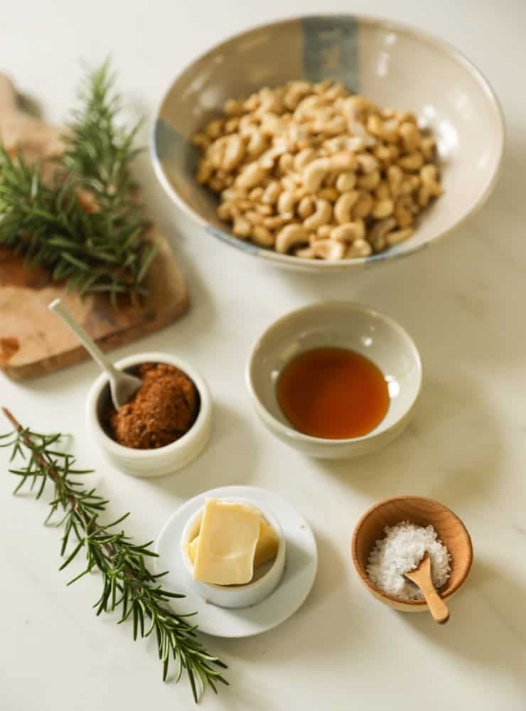 A bowl of raw cashews, honey, butter salt, brown sugar and rosemary on a counter.