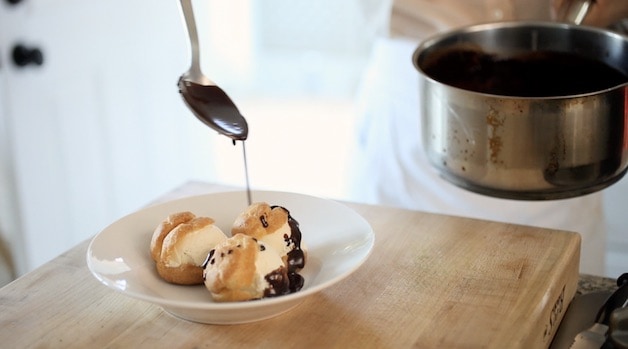 Drizzling chocolate sauce on to 3 profiteroles in a white bowl