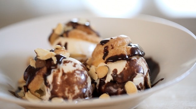 2 profiteroles filled with ice cream and topped with chocolate sauce in a white bowl
