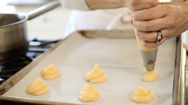 Piping profiterole dough out of a pastry bag onto baking sheets lined with parchment paper