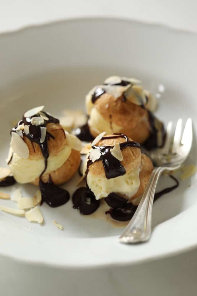 Choux Pastry filled with ice cream and topped with chocolate sauce and toasted almonds in a white bowl.