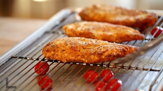 Fried chicken cutlets resting on a baking sheet with a roasting rack with cherry tomatoes below