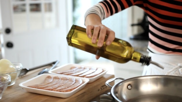 Pouring oil into a pan with two packages of raw sausages off to the side on a cutting board