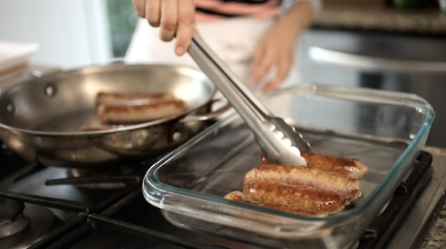 Placing browned sausages from a skillet into a casserole dish with tongs