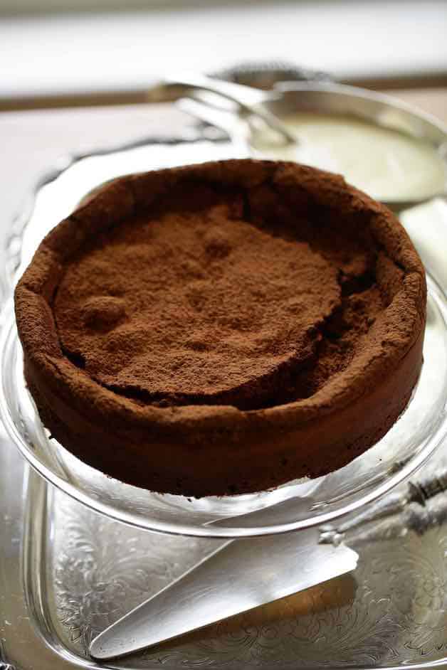 Vertical image of a flourless chocolate cake on a silver tray with silver cake server