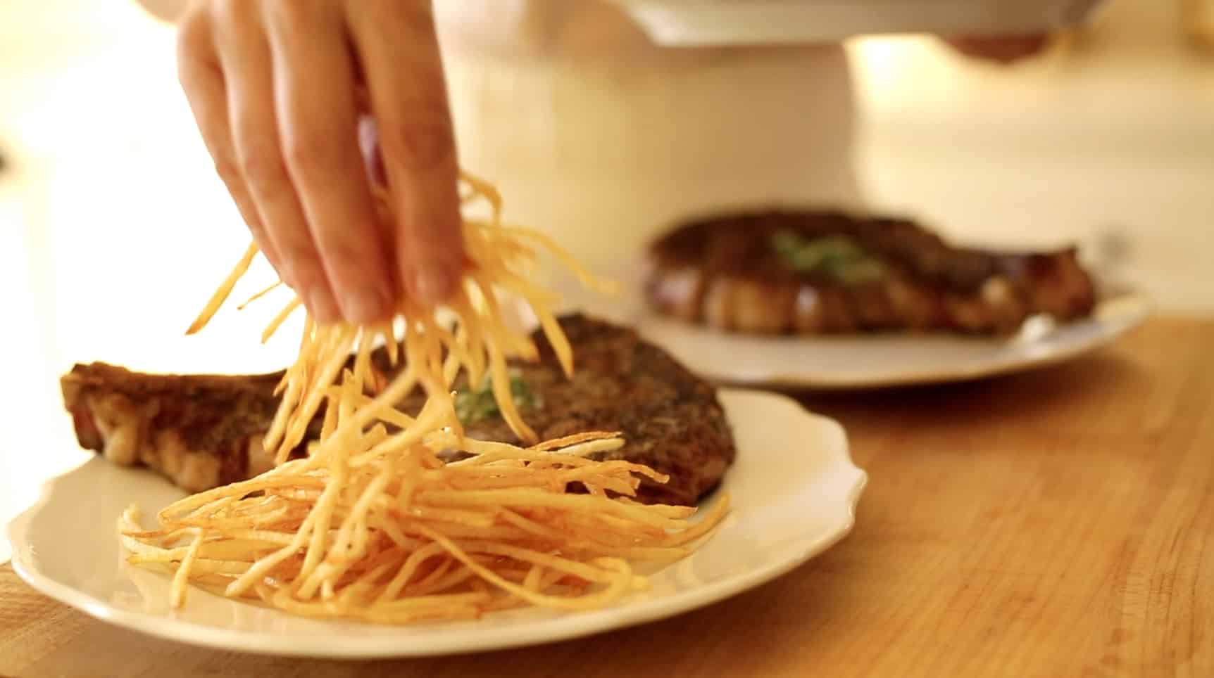 a hand placing a mound of fried french fries on a plate next to a rib-eye steak topped with herb butter