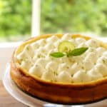 Vertical Image of a key lime pie cheesecake resting on a glass cake stand garnished with a fresh citrus blossom and lime wheel