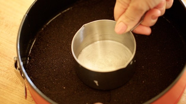 patting down chocolate cookie crumb crust with a stainless steel measuring cup
