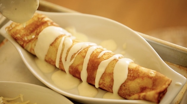 a rolled crepe in a white gratin dish with bechamel sauce drizzled on top