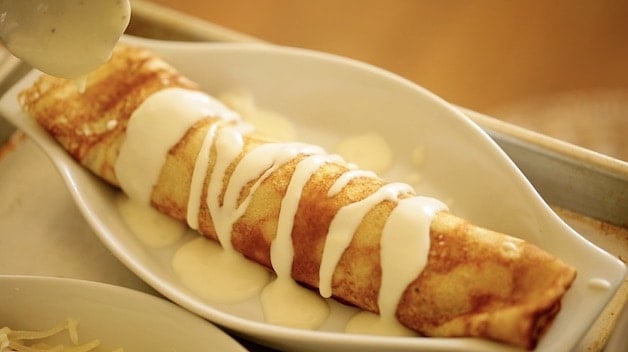 a rolled crepe in a white gratin dish with bechamel sauce drizzled on top