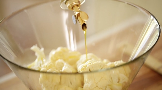 Drizzling olive oil onto raw cauliflower in a glass bowl