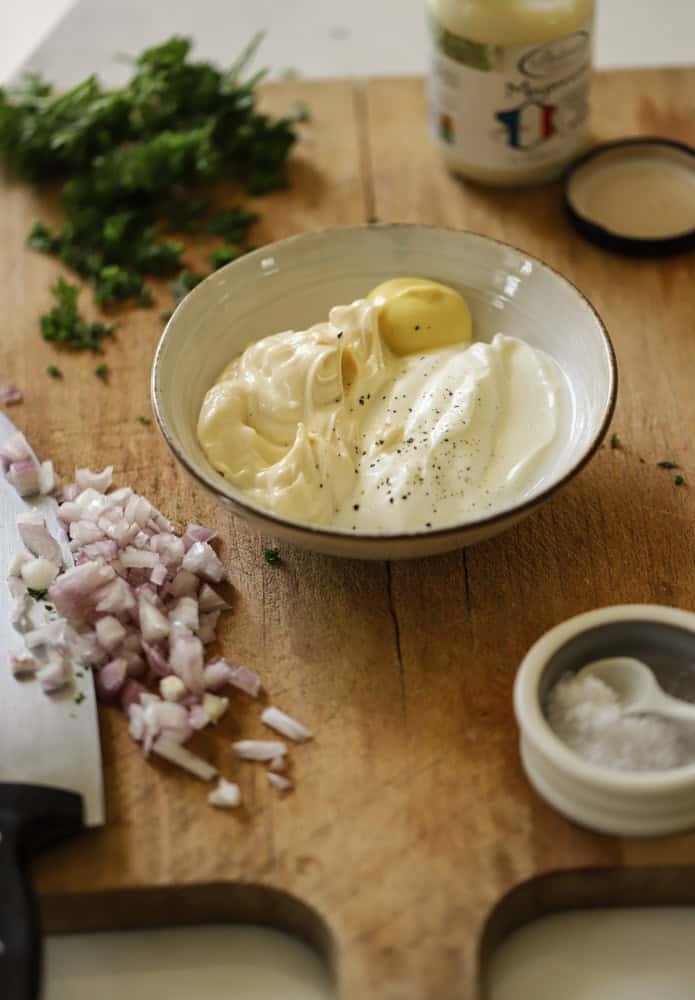 A cutting board with a bowl in the center holding sour cream, mayonnaise and mustard. Herbs and red onion chopped to the side. 