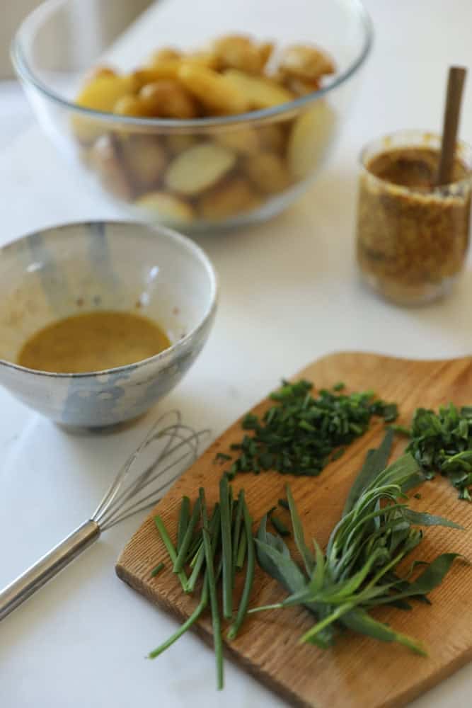 A cutting board with herbs, and a bowl of vinaigrette with a bowl of sliced roasted potatoes.