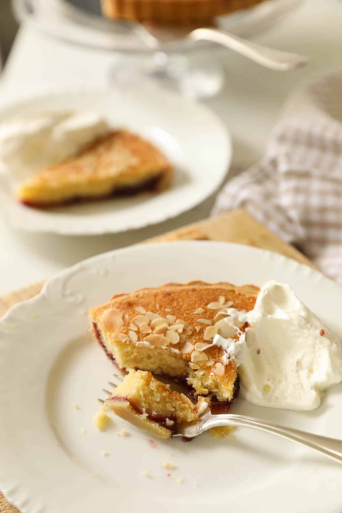 a slice of a cherry bakewell tart with a bite taken out of it, on a white plate with whipped cream.