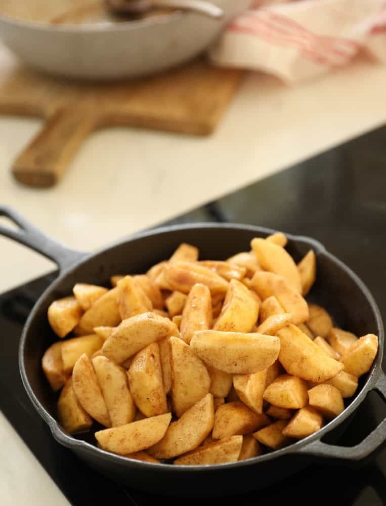 Appl slices tossed in spices, sugar and lemon juice simmering on a cooktop in a cast-iron skillet.