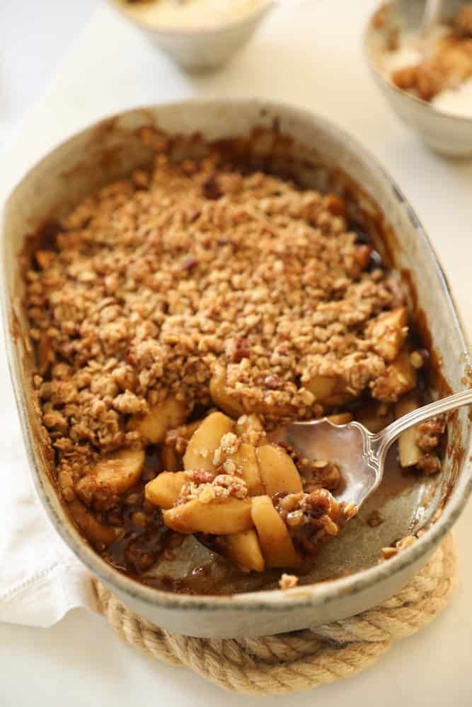 An apple crumble with a serving spoon in it and serving bowls in the distance.