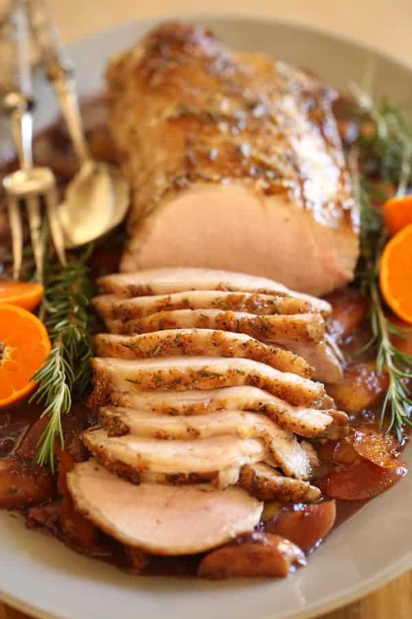 A vertical Image of a Pork loin roast recipe carved on a platter with oranges and rosemary