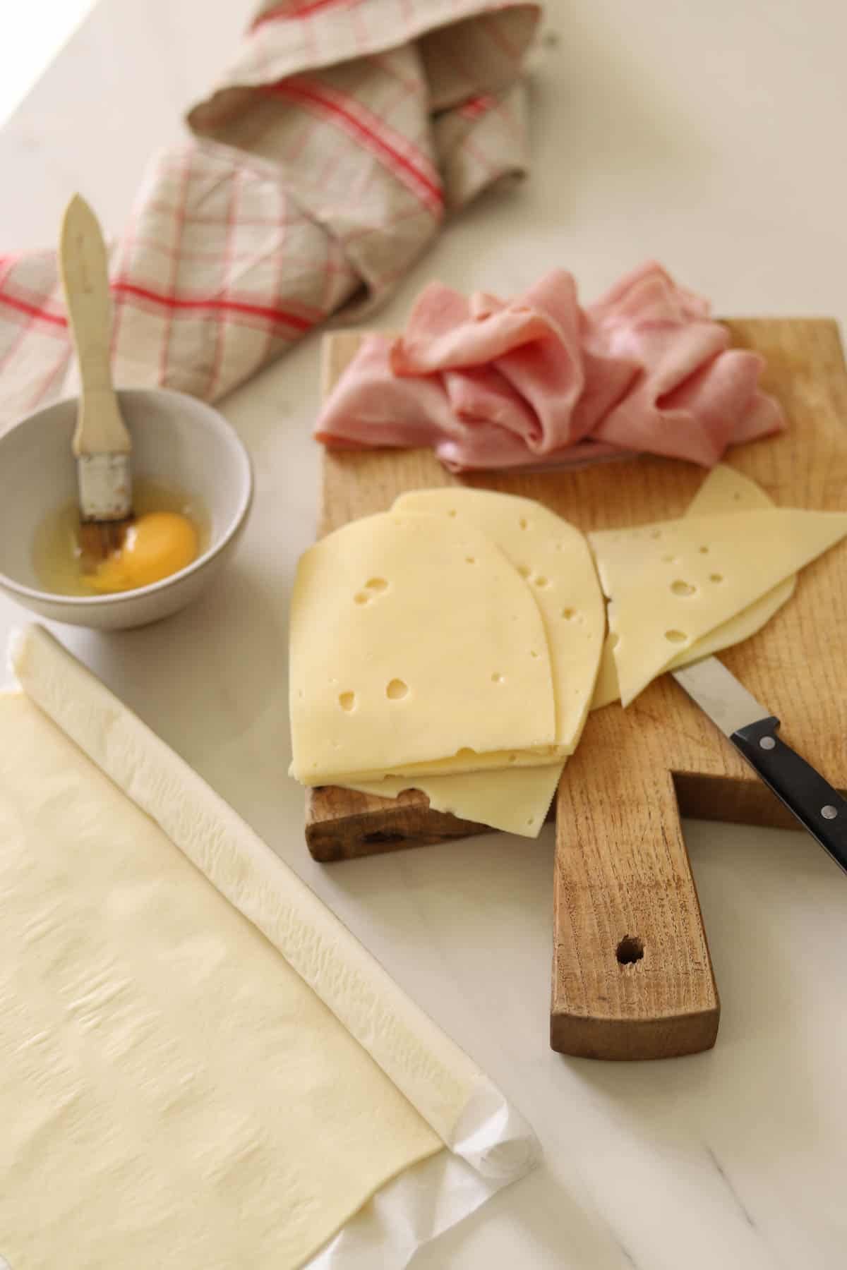 Ham and cheese on a cutting board with puffed pastry and egg in a bowl.