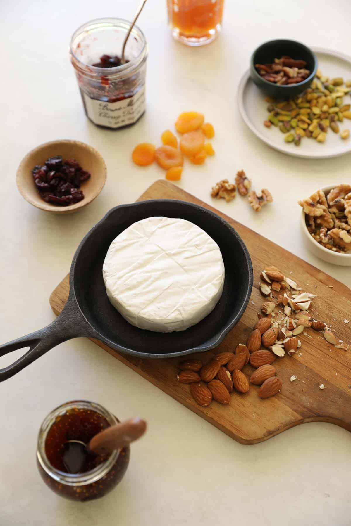 a counter with a skillet of brie showing the rind with jams and nuts