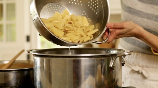 adding cooked rigatoni from a colander to a large pot
