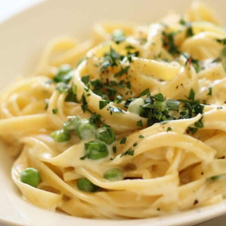 A bowl of pasta in a white bowl with peas and parsley