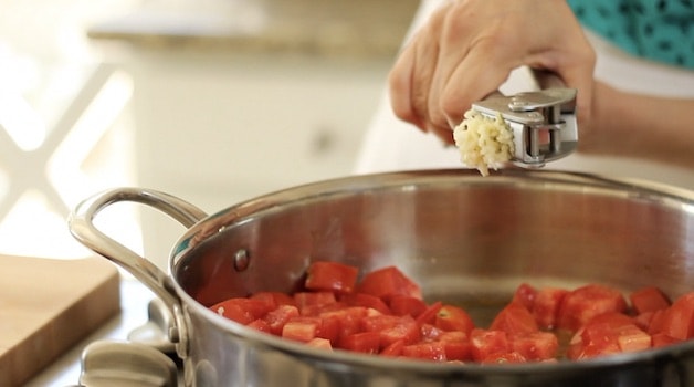Adding garlic from a garlic press to tomatoes in a skillet