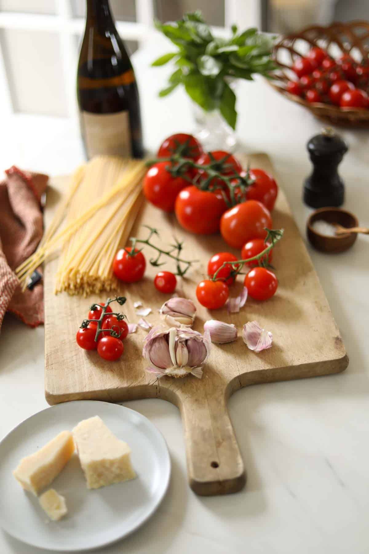 Tomatoes, garlic raw pasta, salt and pepper and parmesan cheese laid out on a cutting board.