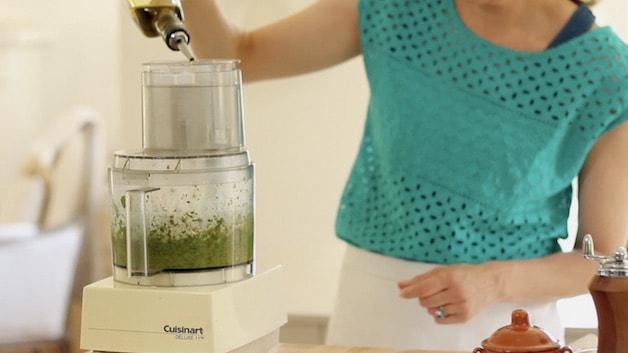 adding olive oil to a food processor filled with basil
