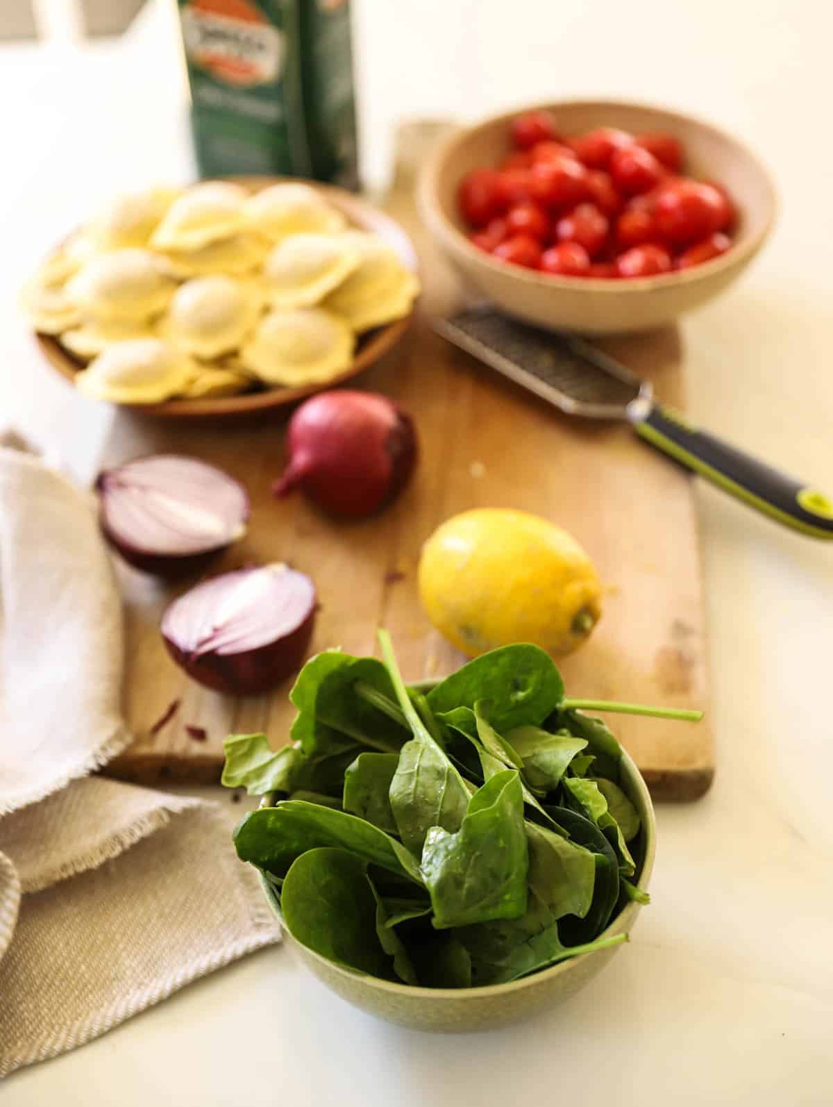Spinach Ravioli, Cherry Tomatoes, Fresh Spinach, Lemon and Red onions laid out on a cutting board.