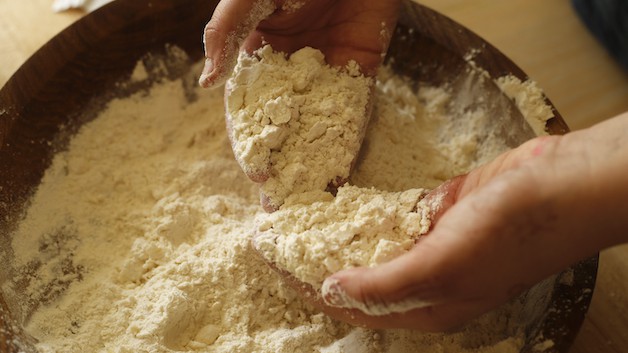 Hands in a wooden bowl of flour and butter mixture creating a coarse meal