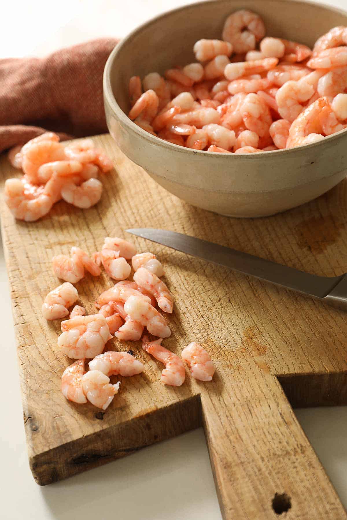 Small cooked shrimp being chopped on a cutting board.