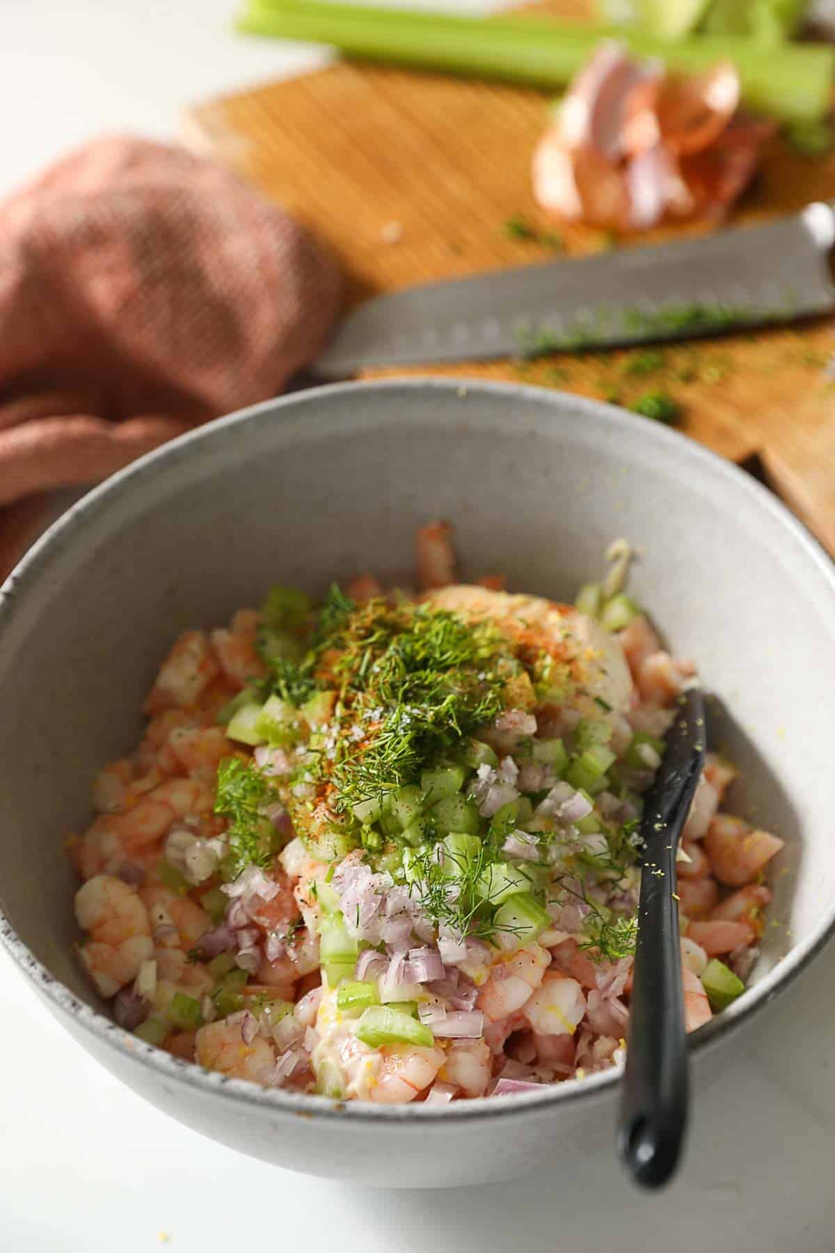 a bowl filled with ingredients like cooked shrimp, celery, herbs and mayonnaise.