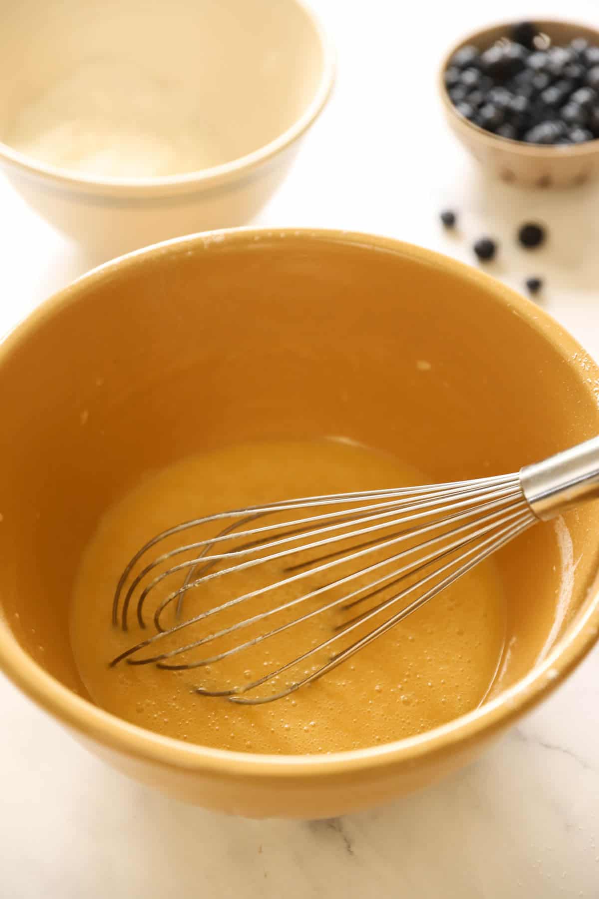 a bowl of batter and a whisk with a smaller dry ingredient bowl in background with a berry bowl