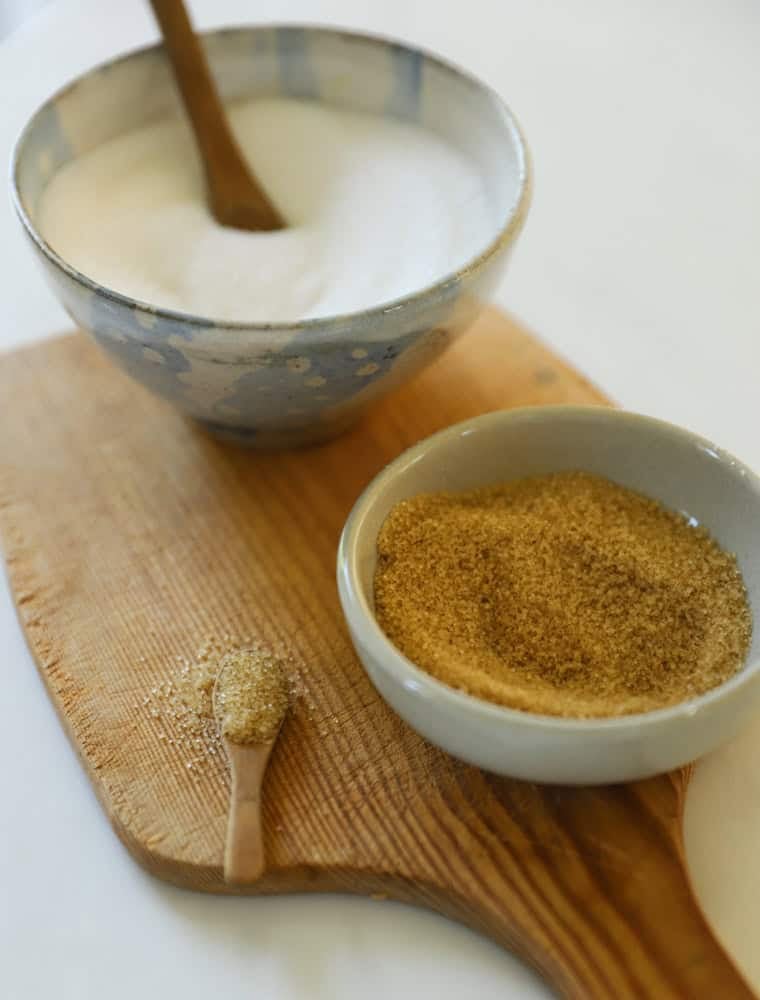 A bowl of white sugar and a bowl of brown sugar on a cutting board.