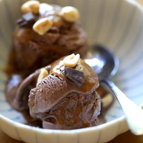 a vertical image of chocolate ice cream scoops and peanut butter sauce with a garnish of nuts and chocolate chunks