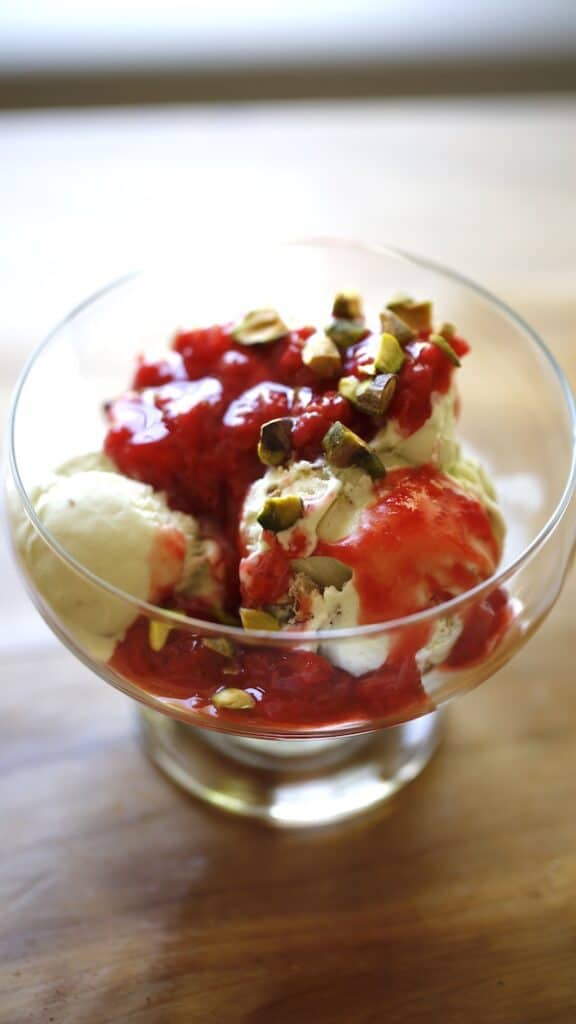 a vertical image of a glass footed ice cream dish with 2 scoops of pistachio ice cream topped with strawberry sauce topping