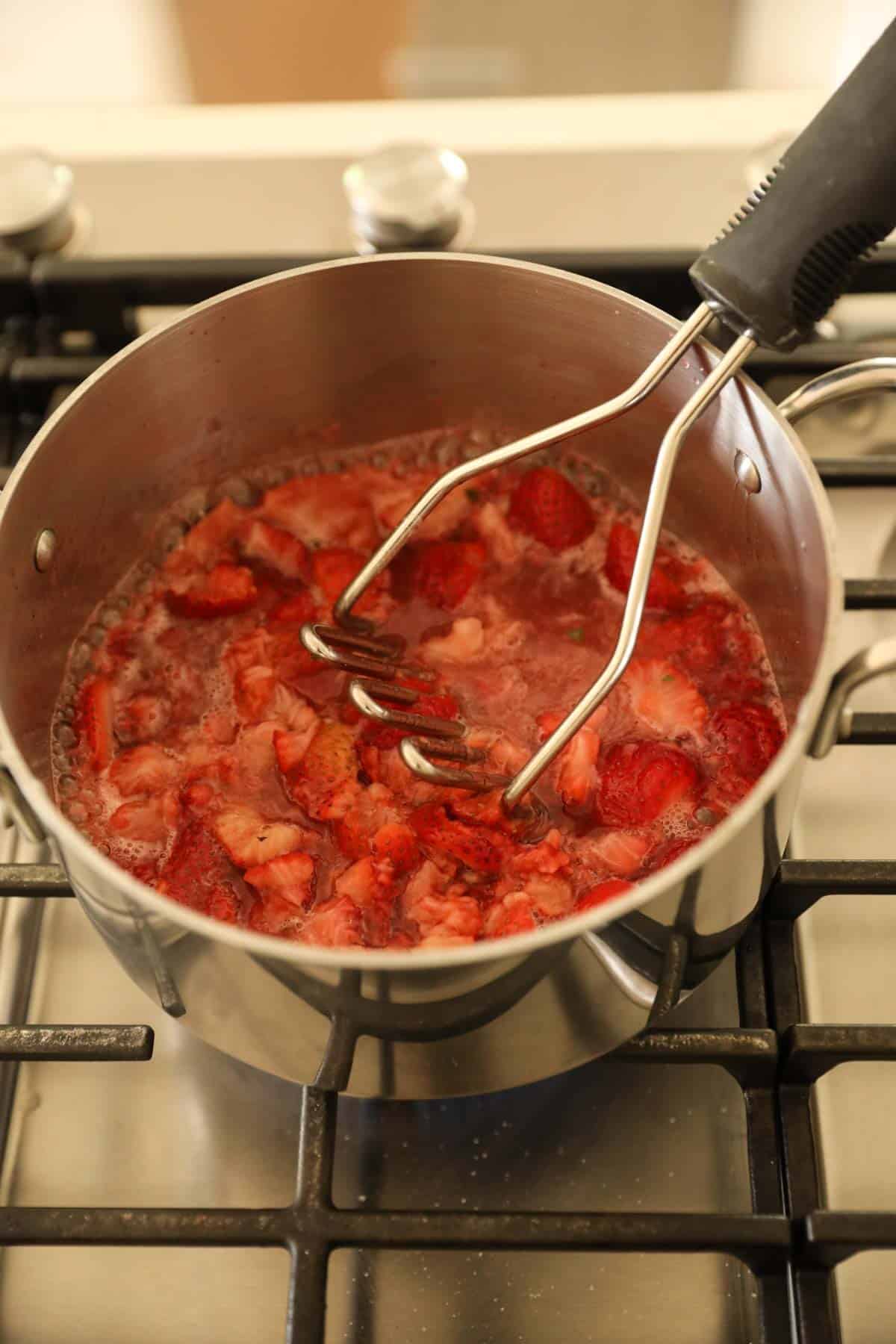 crushing simmering strawberries on a pot with a potato masher