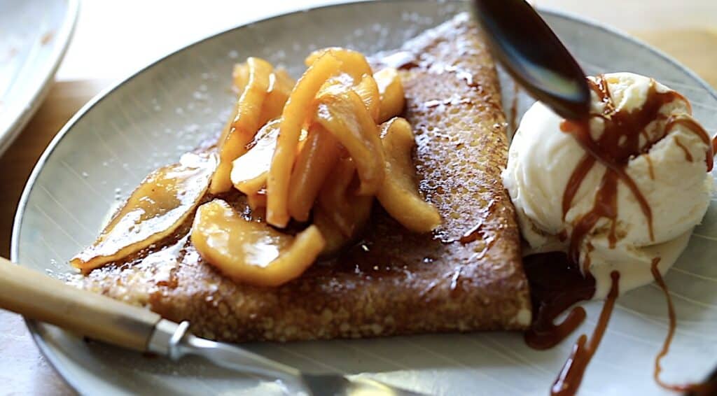 An apple crepe on a plate with a scoop of ice cream being drizzled with caramel sauce