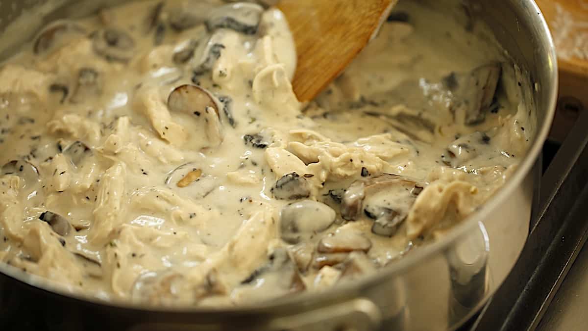 a skillet of bechamel sauce with mushrooms, chicken and a wooden spoon