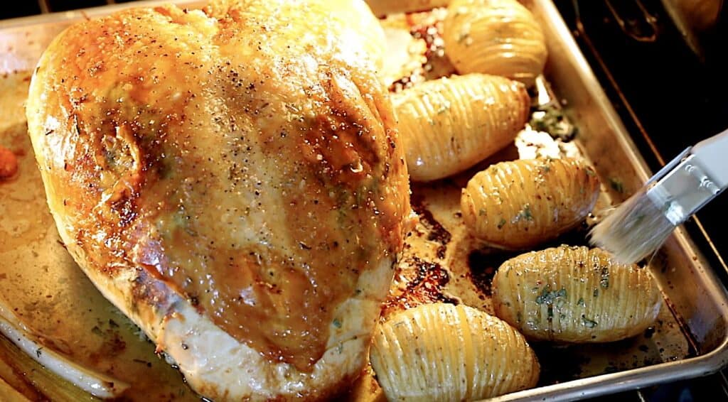 close up of a sheet pan of turkey roasting and a pastry brush brushing sage butter on potatoes beside it