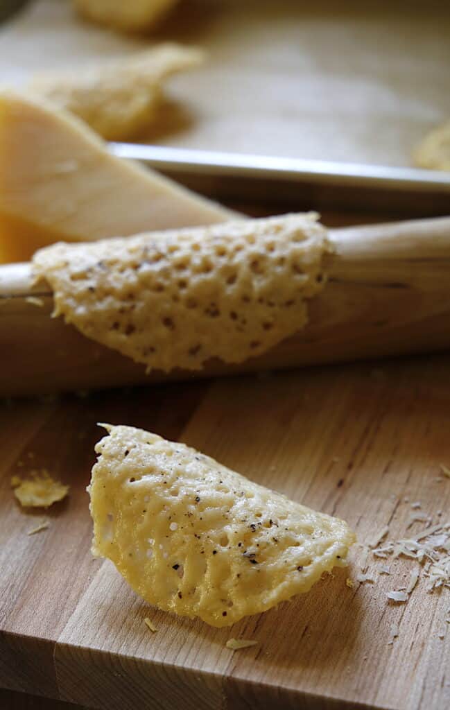 a vertical image of a parmesan tuile on a cuttng board with a tuille around a rolling pin on background