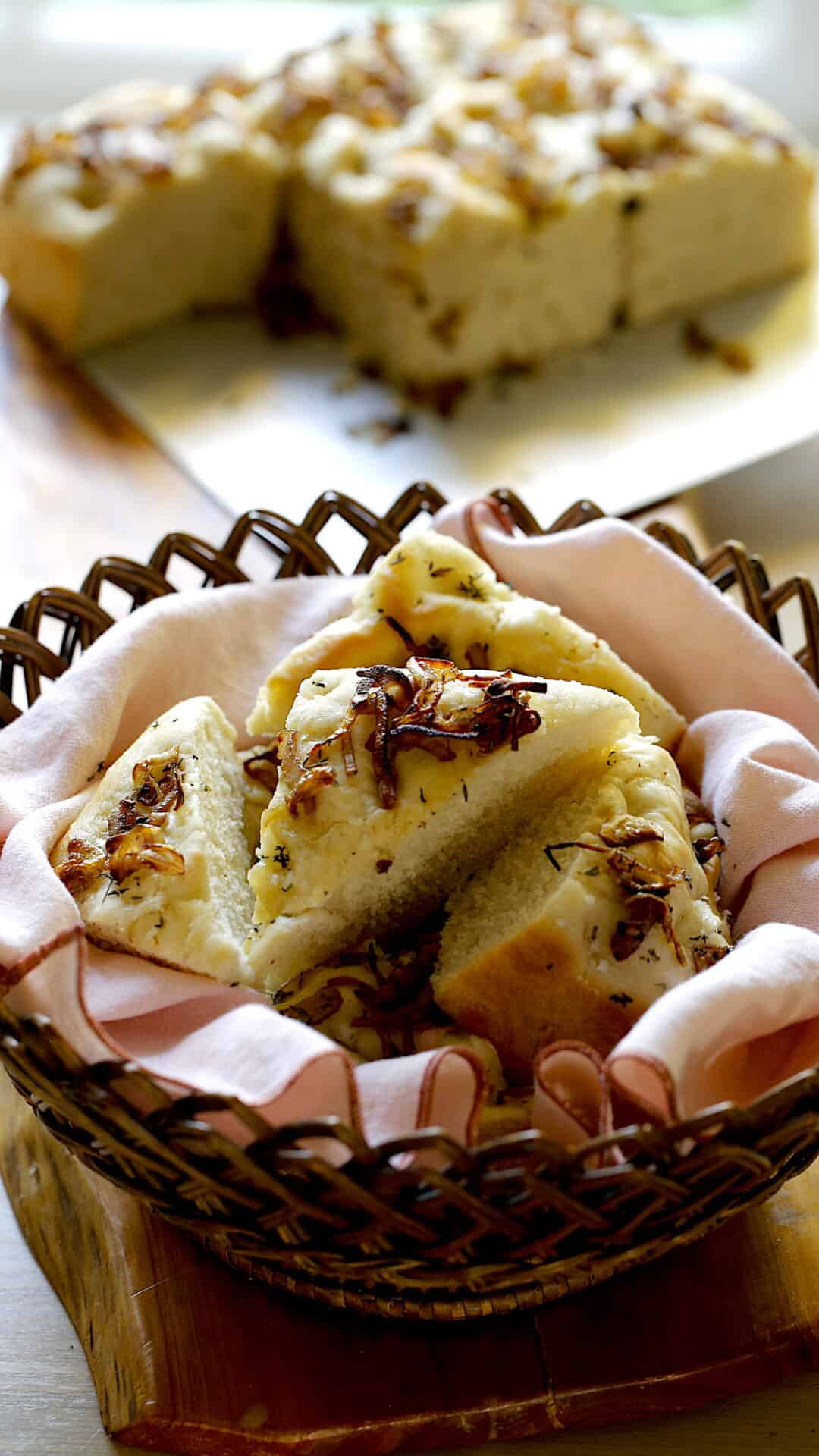 A vertical image of a bread basket filled with a Focaccia bread slices with a pan of focaccia bread in the background