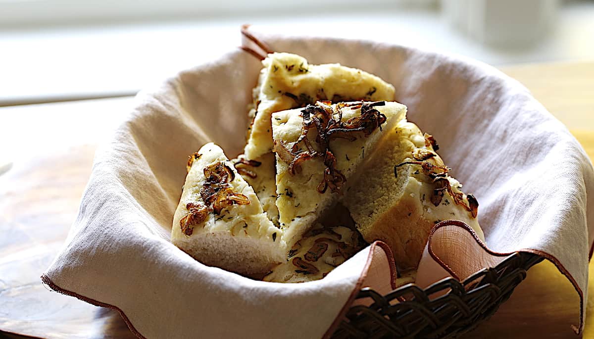 A bread basket filled with slice focaccia bread topped with shallots