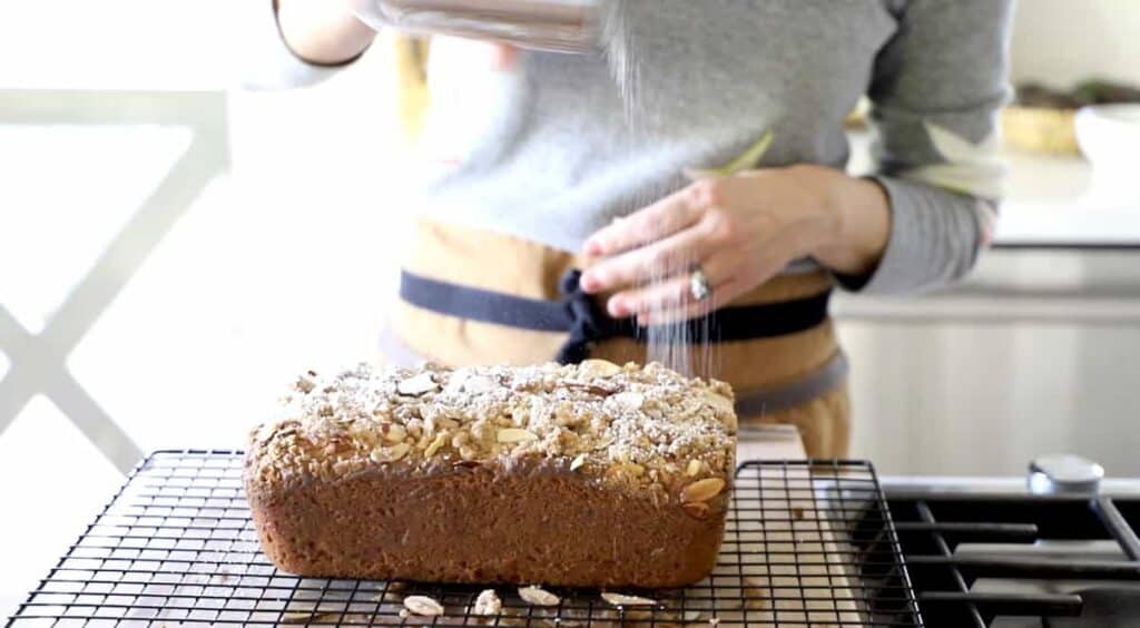 A person dusting a crumb cake loaf with powdered sugar