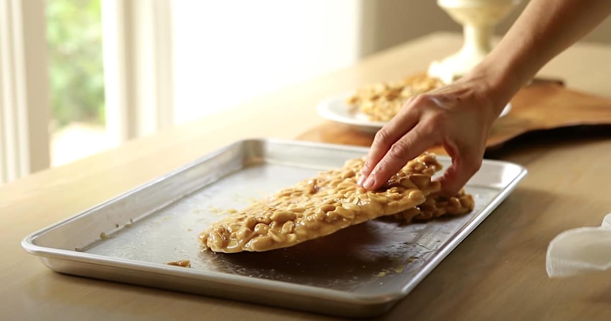 a person lifting up a mass of hardened peanut brittle on a sheet pan