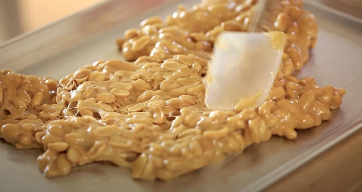 a spatula pressing down hot peanut brittle onto a sheet pan