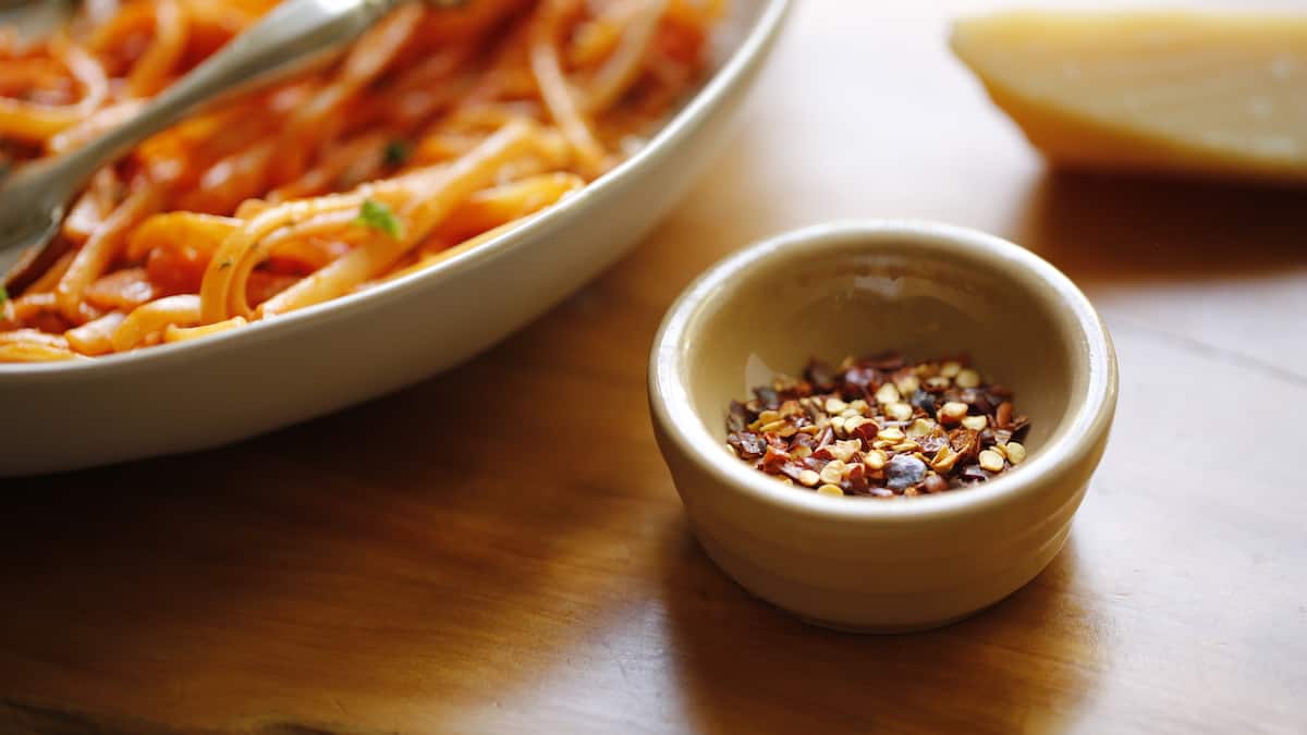 a little bowl of red pepper flakes with a bowl of pasta and a wedge of parmesan cheese in the background